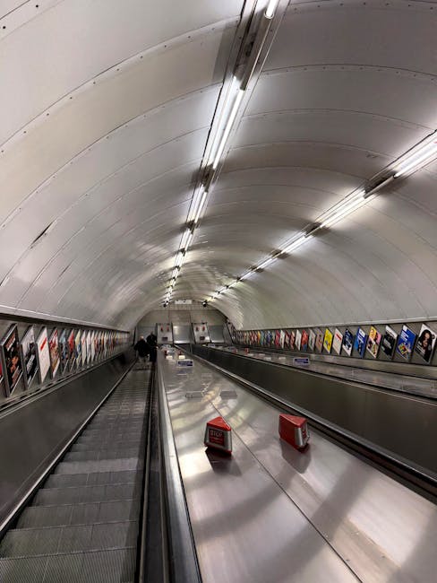 A long, curved underground passageway with smooth, white arched walls and ceiling illuminated by evenly spaced fluorescent strip lights. On the left side, there is a moving walkway with a metal surfac