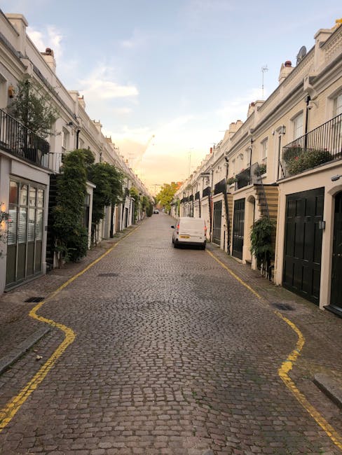 A narrow cobblestone street in a residential area with white terraced houses on both sides, each featuring black or brown garage doors and small balconies with potted plants. The street is lined with 