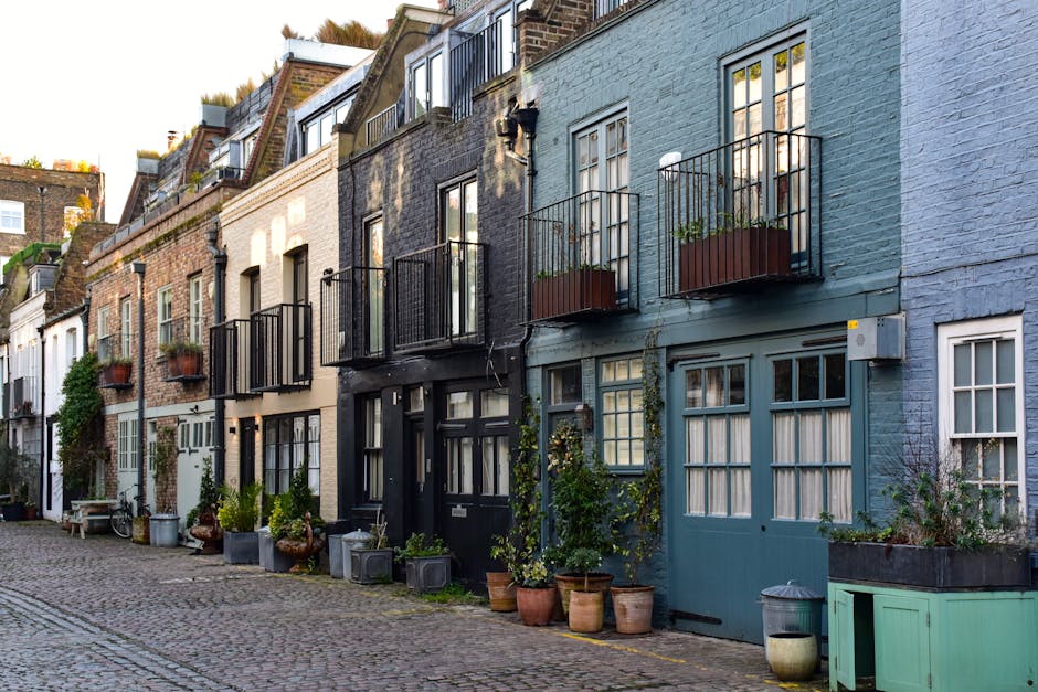 A narrow cobblestone street in a residential area with white terraced houses on both sides, each featuring black or brown garage doors and small balconies with potted plants. The street is lined with 